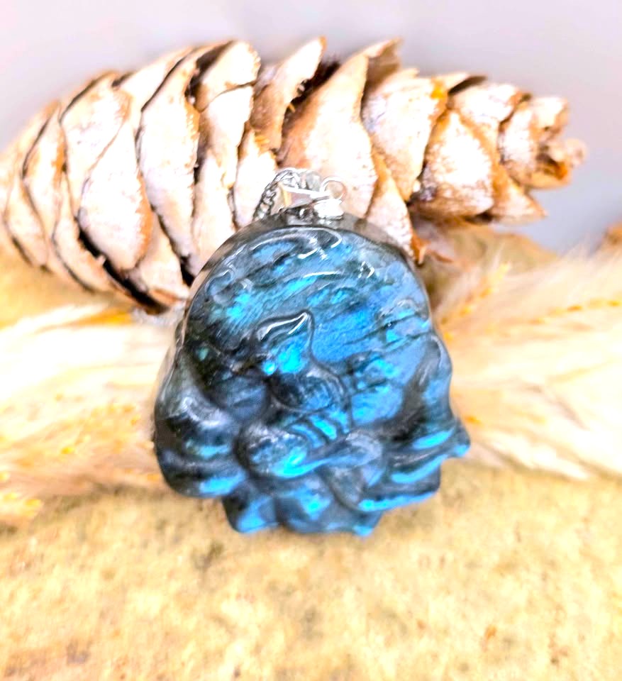 A carved blue labradorite pendant with a silver bail, resting on a pine cone and dried grass against a neutral background.
