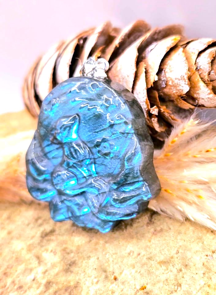 A carved blue pendant with a silver bail, resting on a sandy surface in front of a pine cone and dried grass.
