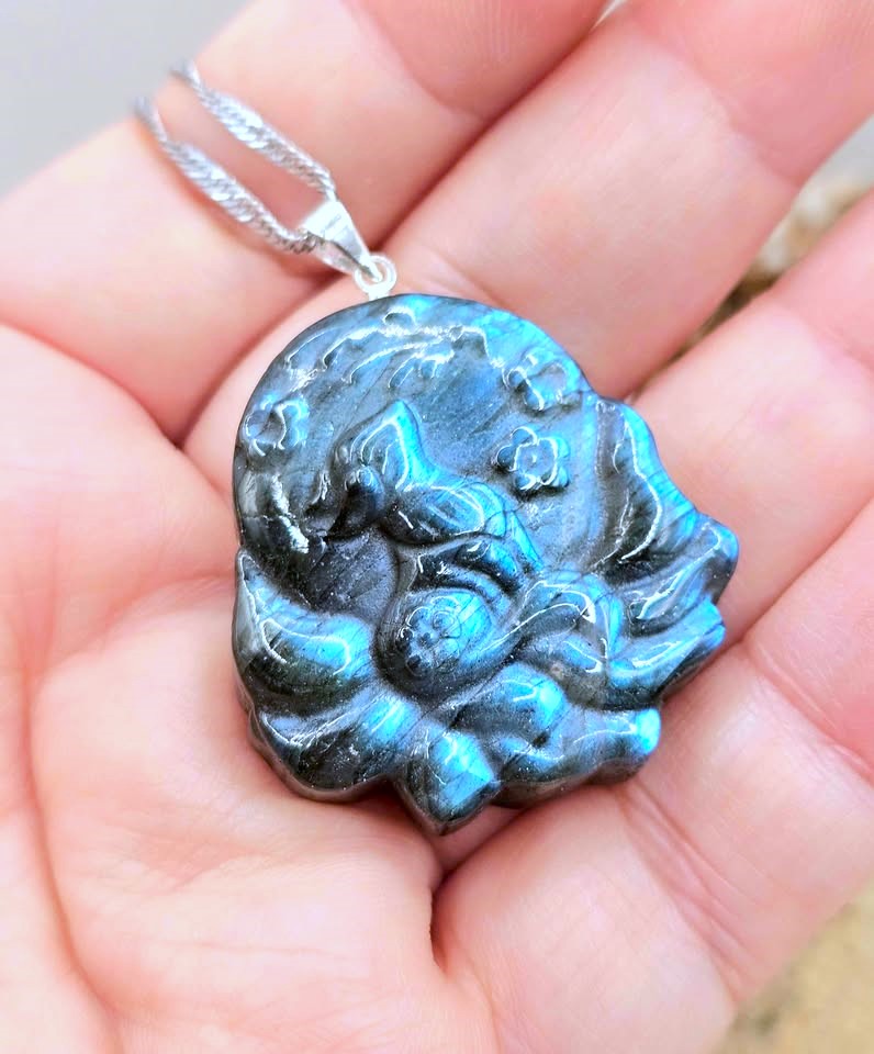 A close-up of a hand holding a carved pendant necklace. The pendant is made of a dark, textured material with a bright blue-green iridescent sheen, suspended from a silver chain.