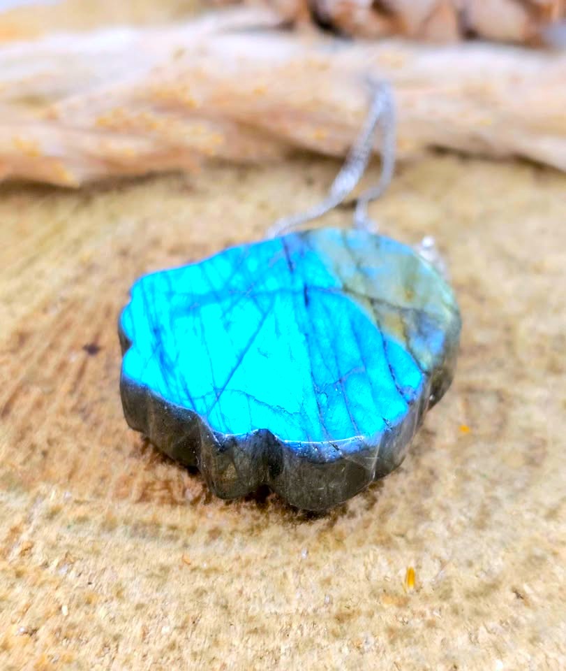 A handcrafted irregular-shaped pendant made from a polished labradorite stone showing vibrant blue flashes and dark gray edges, suspended from a silver wire on a textured sandy surface.