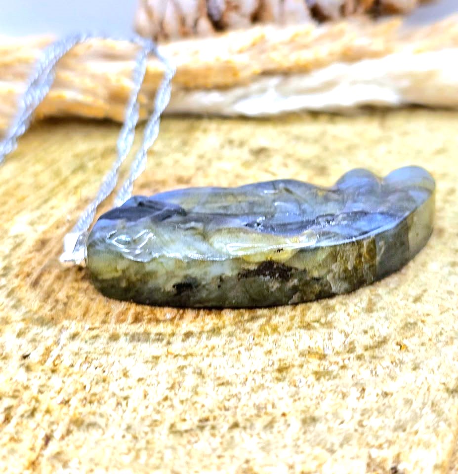 A raw, polished Labradorite pendant necklace resting on a textured stone surface, showing natural blue, gray, and green color variations.