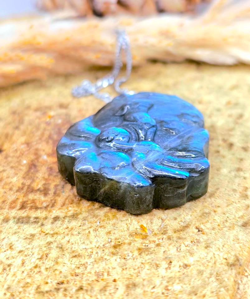 A close-up of a carved black stone pendant with bright blue iridescent flashes, hanging from a silver-toned chain against a sandy, natural background.