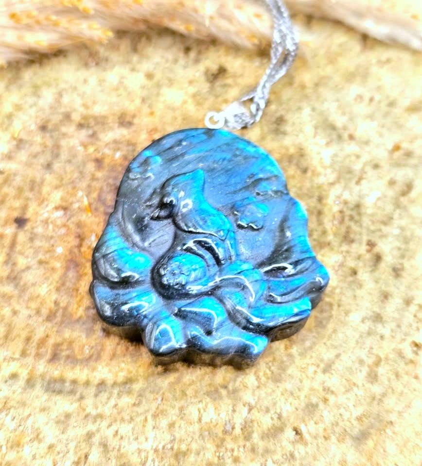 A close-up of a carved stone pendant with a silver chain, featuring a highly polished blue and black iridescent surface, resting on a textured tan stone background.