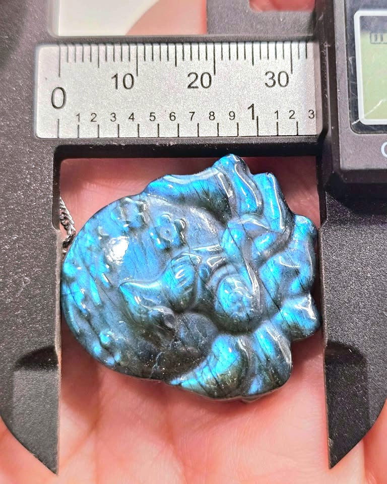 A close-up of a carved labradorite pendant held in a hand under a metal caliper. The pendant has a blue-green iridescent labradorite finish with dark gray to black natural inclusions.