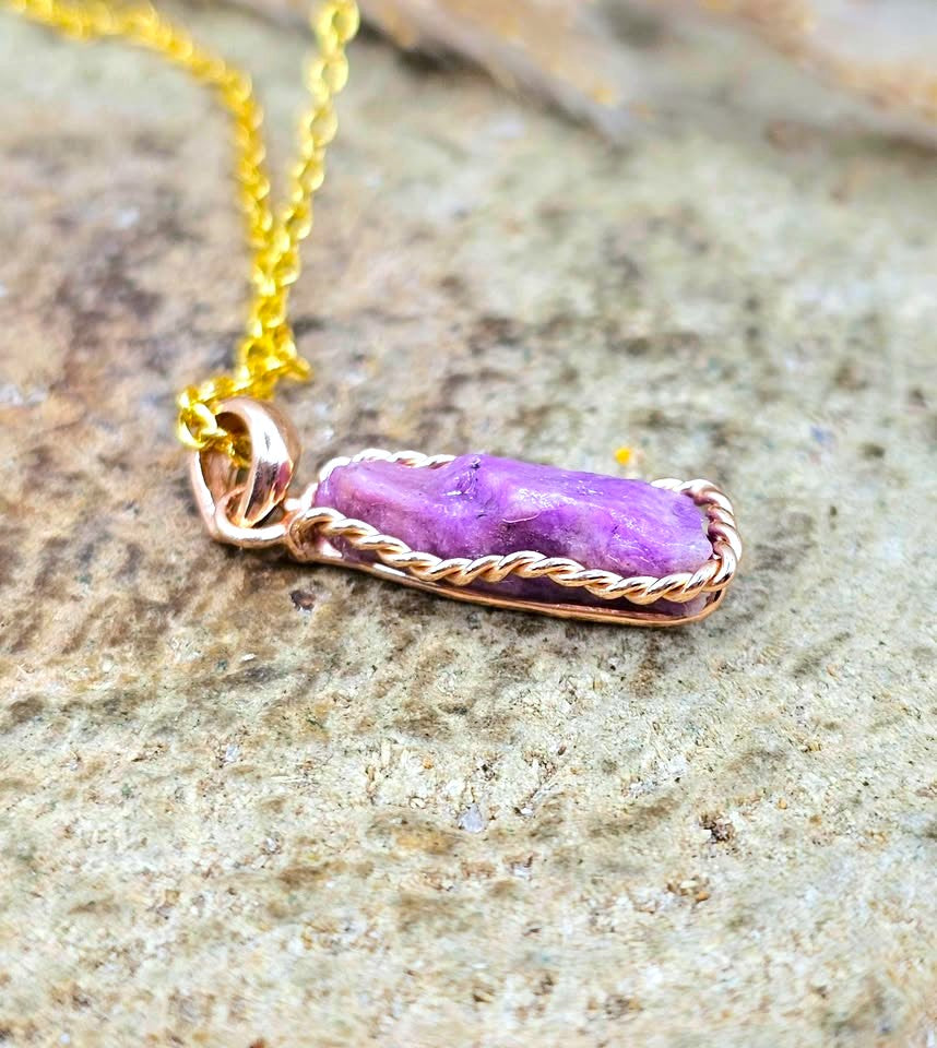 A close-up of a gold chain necklace with a rectangular pendant wrapped in twisted gold wire, holding a polished purple stone, resting on a textured gray stone surface.