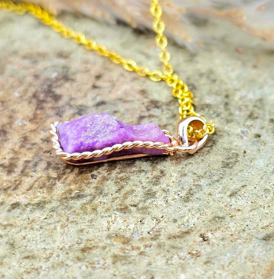 A gold chain necklace with a handmade pendant featuring a rough purple stone wrapped in twisted rose gold wire, resting on a textured gray stone surface.