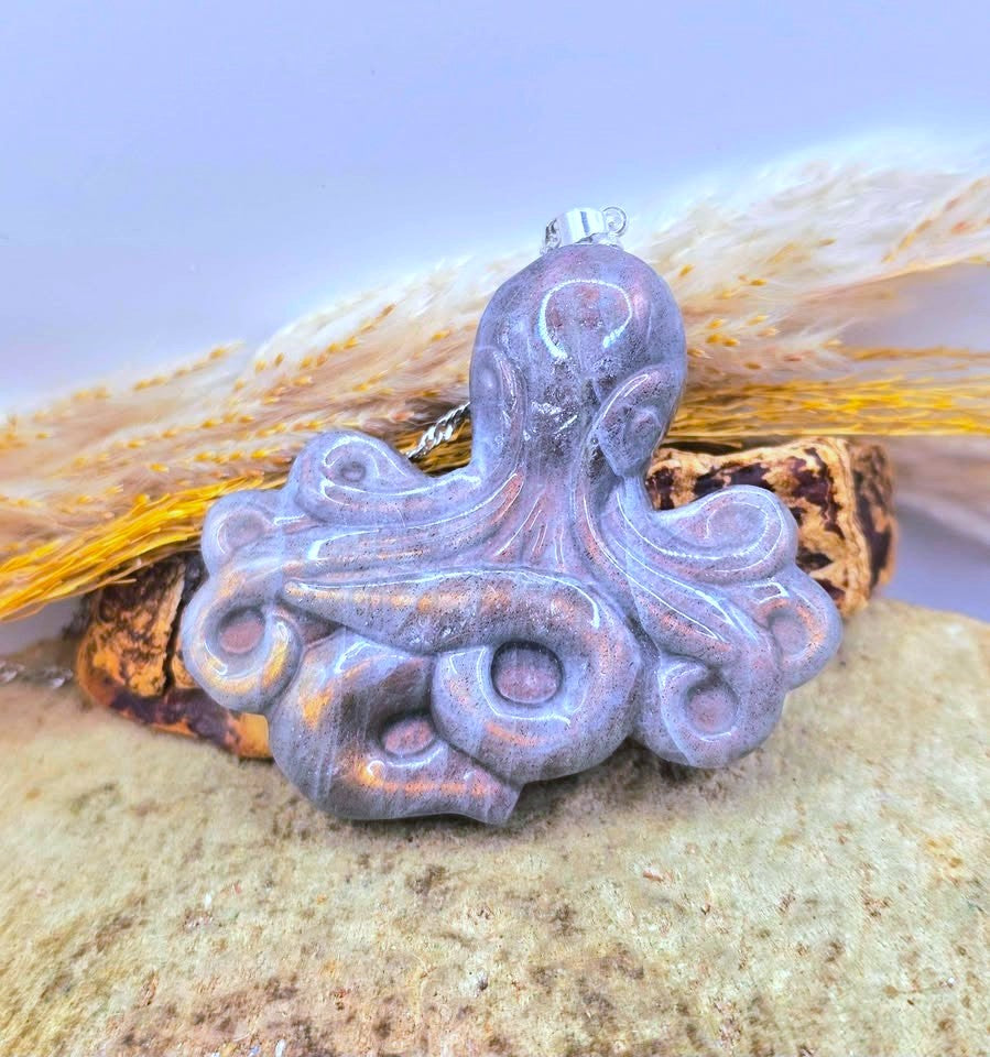 A carved octopus pendant with a glossy lavender-purple finish and subtle coppery highlights, resting on a textured stone with dried grass and wood in the background.