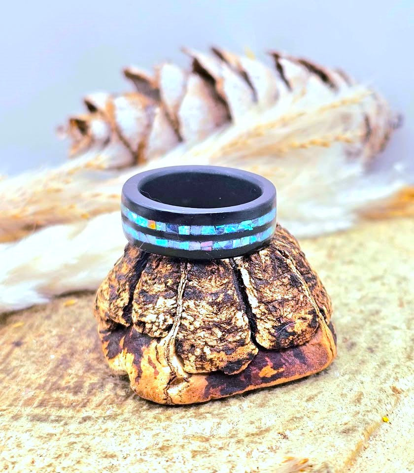 A black wooden ring with a colorful inlay of small invertebrate shell pieces, displayed on a textured brown bark stand with a feather and pinecone in the background.