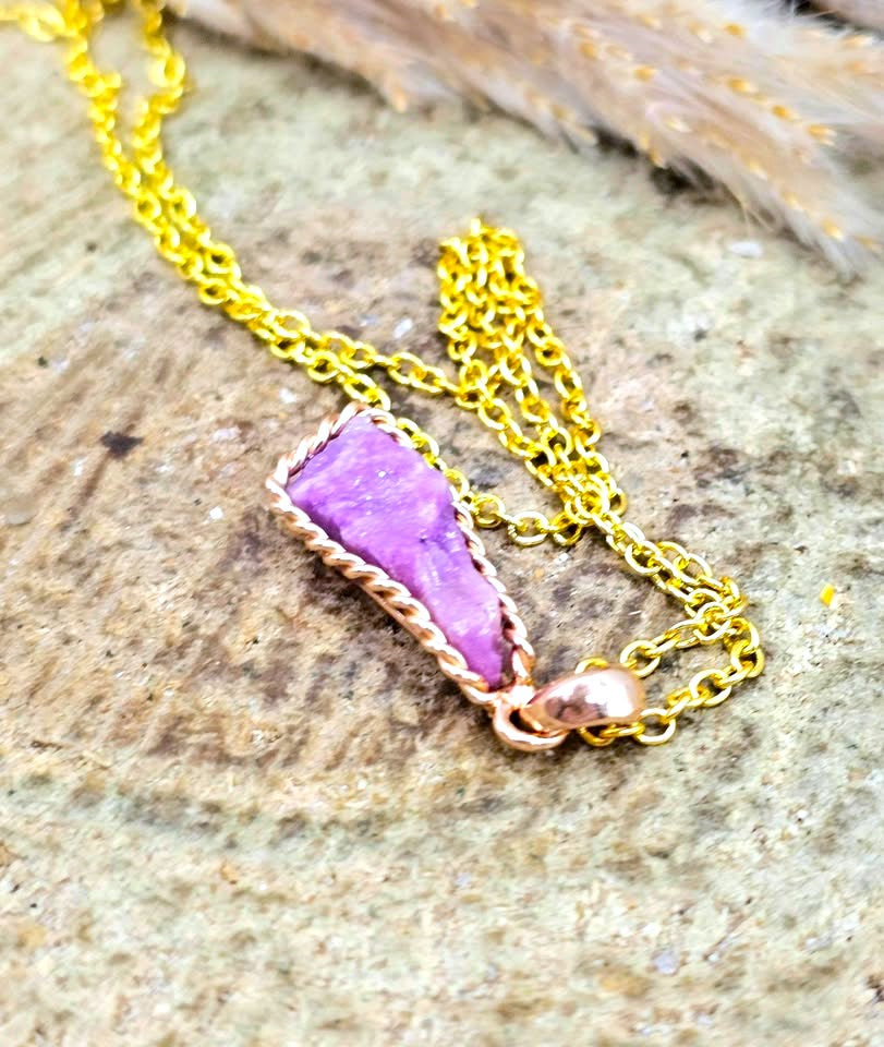 A gold chain necklace featuring a raw purple stone pendant wrapped in a twisted rose gold wire border, resting on a textured stone surface.