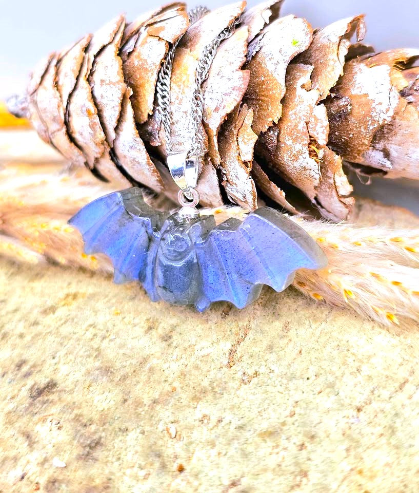 A silver bat pendant necklace with a blue iridescent resin bat wing charm, displayed on a pine cone and dried grass against a sandy background.