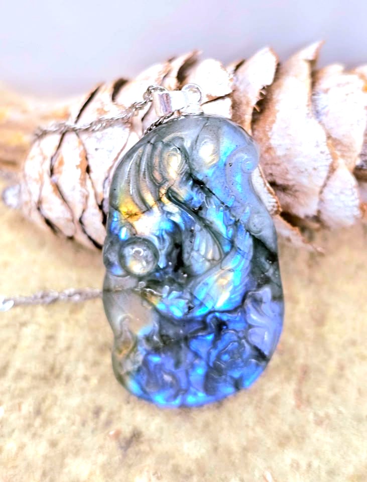 A carved labradorite pendant featuring a detailed figure, resting on a pinecone with a silver chain against a neutral background.