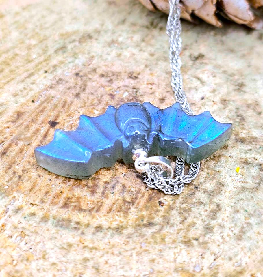 A bat-shaped pendant necklace with a blue iridescent finish and a silver chain, resting on a textured stone surface.