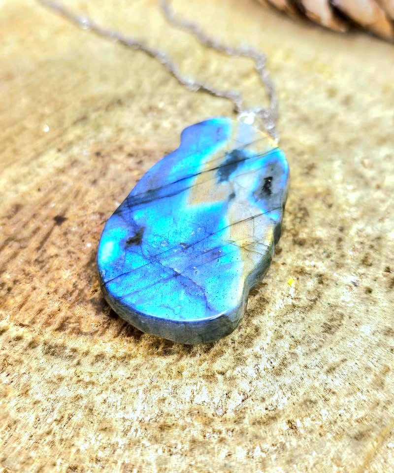 A teardrop-shaped labradorite gemstone pendant necklace with vibrant blue and green iridescent flashes, hanging from a silver chain on a textured stone surface.