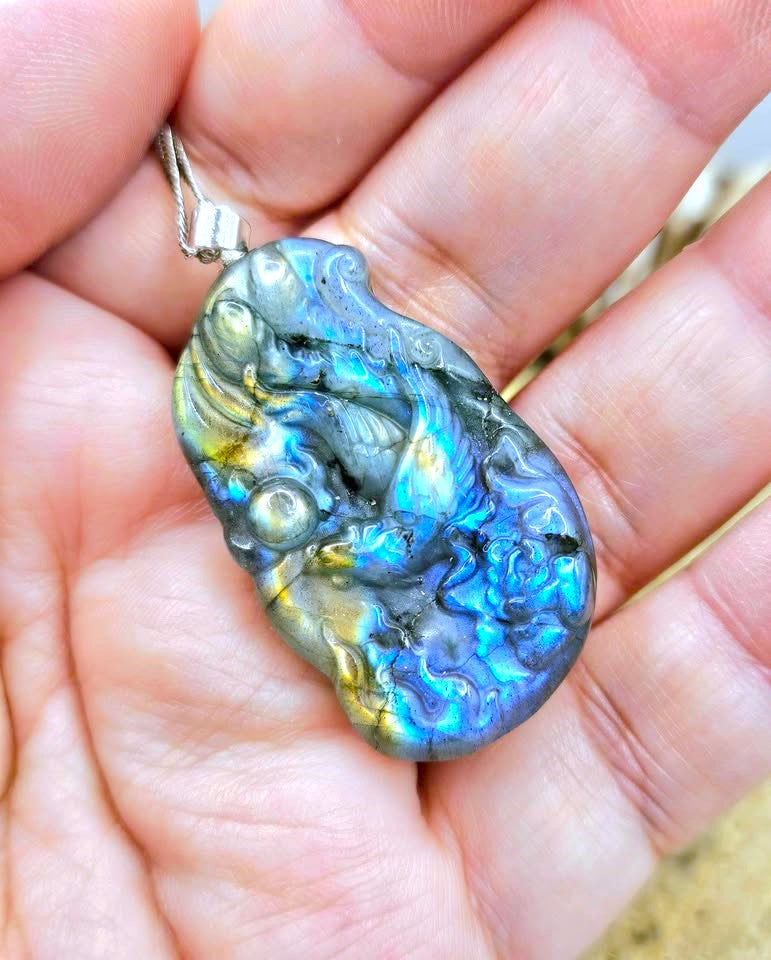 A close-up of a hand holding a polished labradorite pendant with a silver bail. The pendant displays a highly iridescent play-of-color with vibrant flashes of blue, green, gold, and gray.