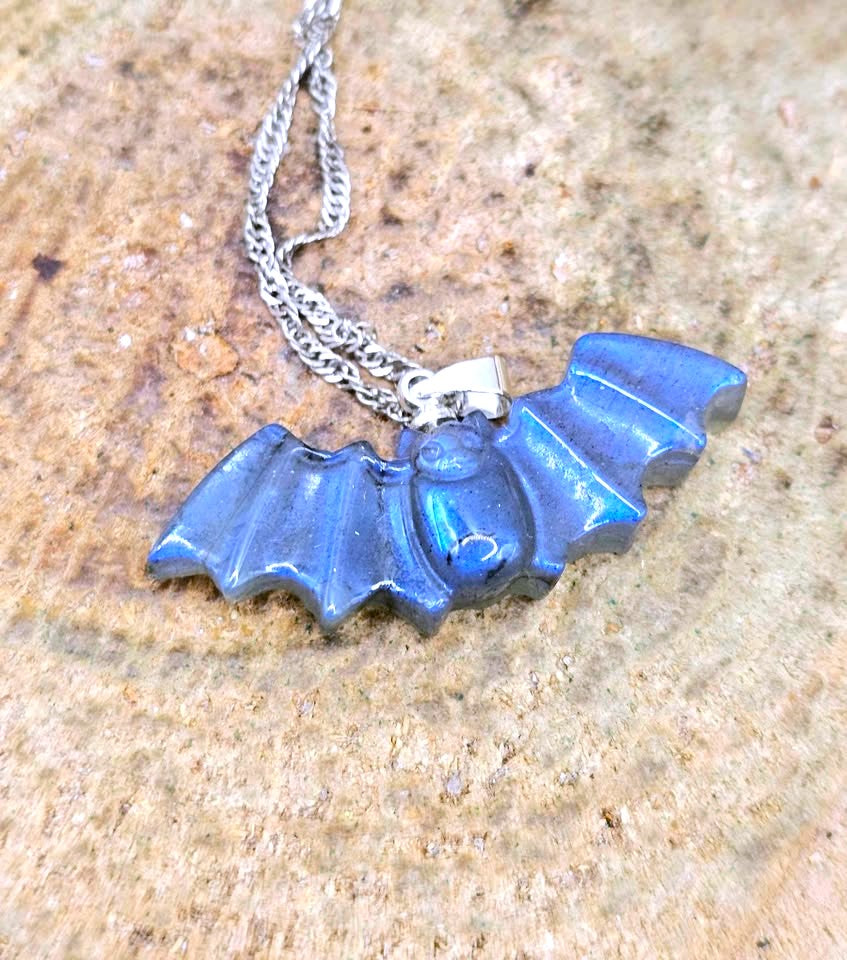A blue bat-shaped pendant necklace with a silver chain, displayed on a textured brown stone surface.