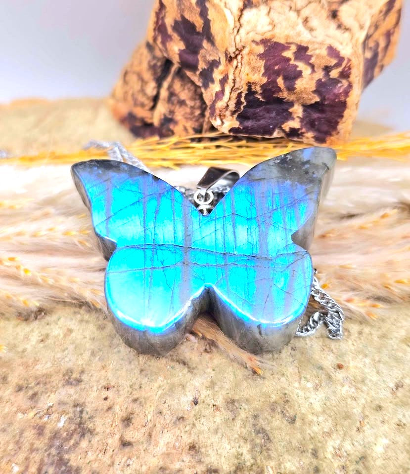 A butterfly-shaped pendant necklace made from iridescent turquoise-blue labradorite with a silver-toned bail and chain, displayed on a textured stone surface with dried grass and a decorative rock in the background.