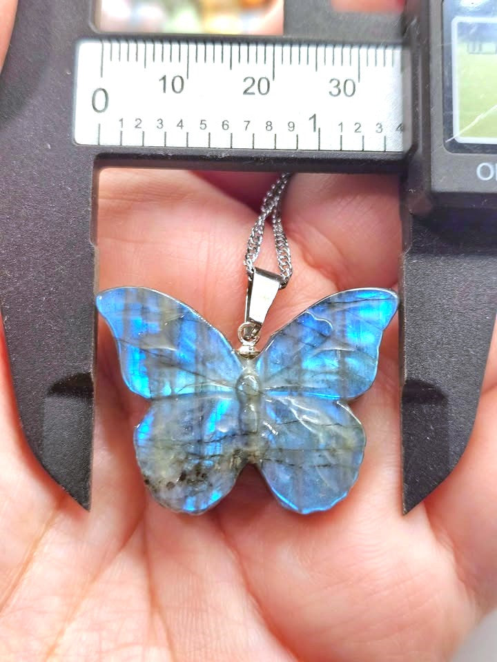 A butterfly-shaped pendant made of iridescent blue labradorite stone with a silver bail and chain, held in a person's hand under a caliper to show its size.