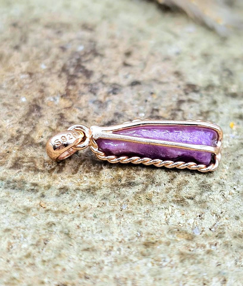 A 925 sterling silver pendant with a purple stone wrapped in twisted rose gold wire, resting on a textured stone surface.