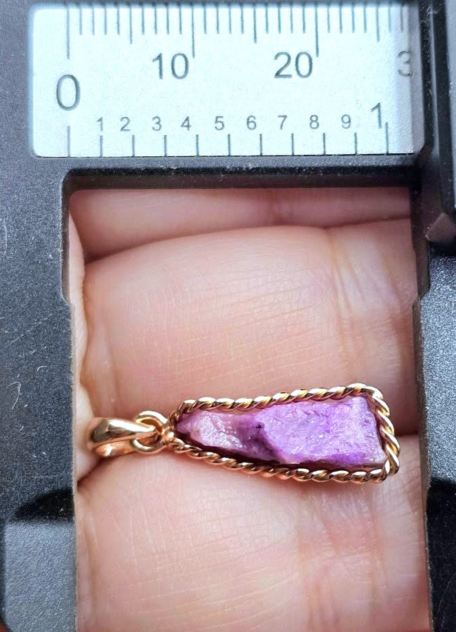 A close-up of a gold pendant with a textured, irregular purple stone, resting on a person's fingers with a metal caliper ruler visible at the top for scale.