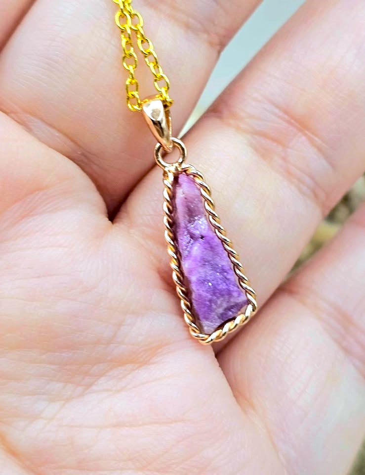 A gold chain necklace with a triangular purple stone pendant, held in a person's hand. The pendant is framed by a twisted gold border.