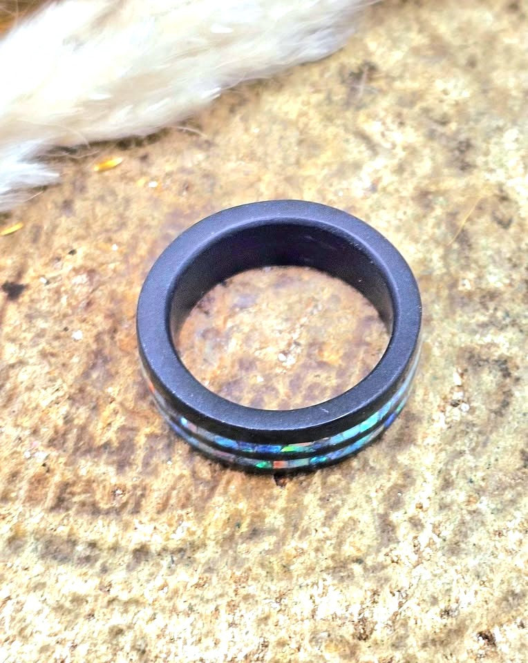 A black ring with a thin inlay of iridescent multicolor opal-like material, resting on a textured brown stone surface.