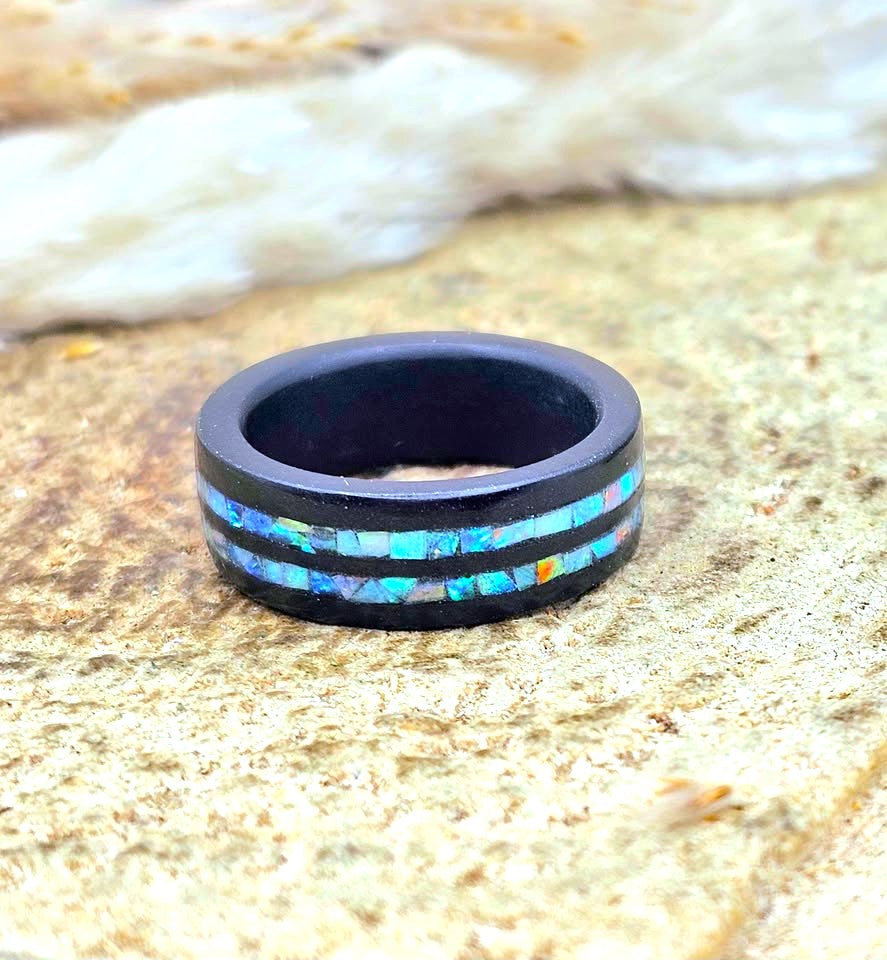 A matte black ring with two inlaid horizontal bands of iridescent blue, green, and orange opal, resting on a sandy surface with blurred white fur in the background.