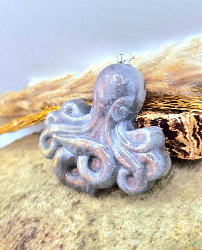 A carved octopus-shaped pendant with a metallic silver finish and a small silver loop at the top, displayed on a textured surface with dried grass and a decorative object in the background.