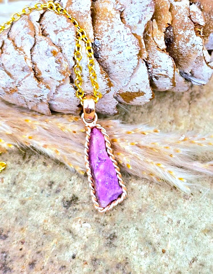 A gold chain necklace with a teardrop-shaped purple stone pendant, set in a twisted gold bezel, displayed on a textured stone surface with a pinecone and dried grass.
