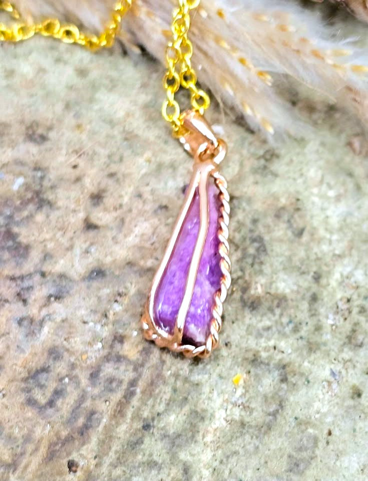 A close-up of a pendant necklace with a purple stone set in a copper-toned twisted wire frame, hanging from a gold-tone chain against a textured stone surface.