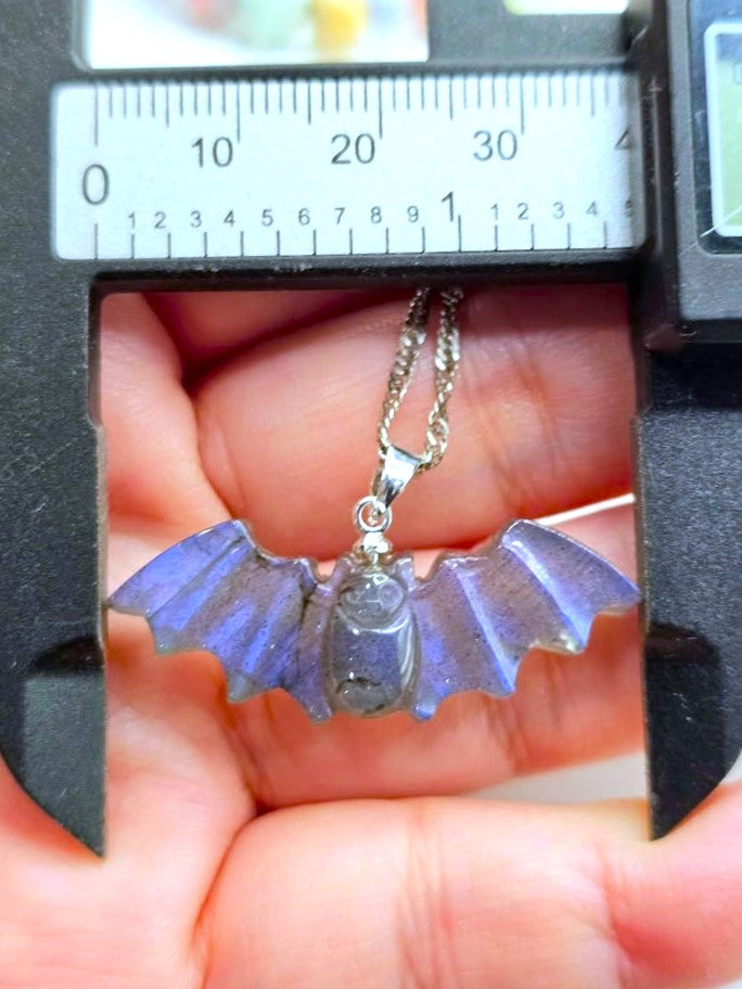 A bat-shaped pendant necklace with purple iridescent wings and a silver-toned chain, held in a hand under a caliper to show its size.
