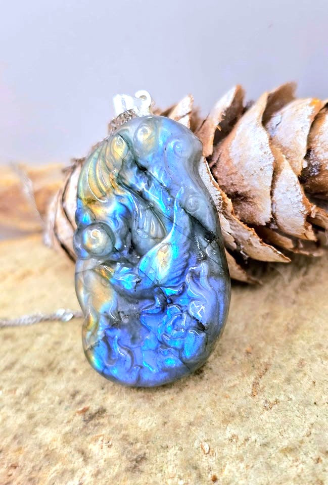 A carved labradorite pendant with an iridescent blue, green, and gold finish, resting on a pine cone with a silver bail at the top.