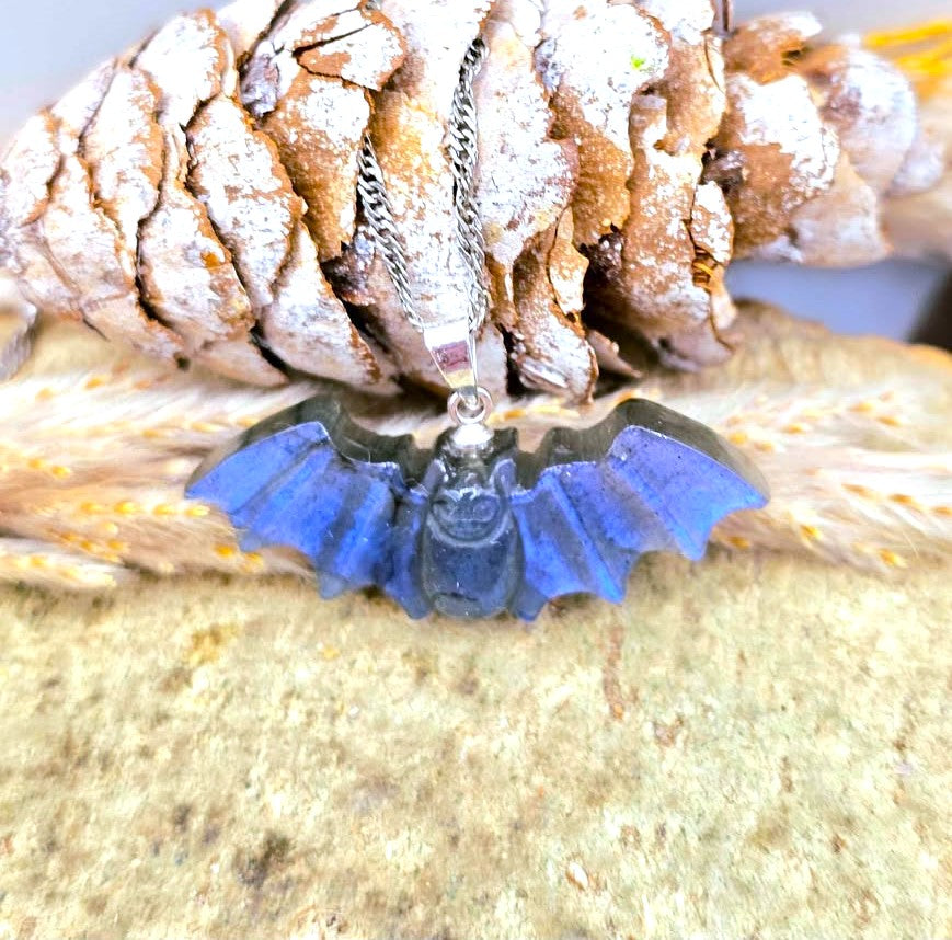 A blue bat pendant necklace with a silver-toned chain, displayed on a textured stone surface with a pine cone and dried grass in the background.