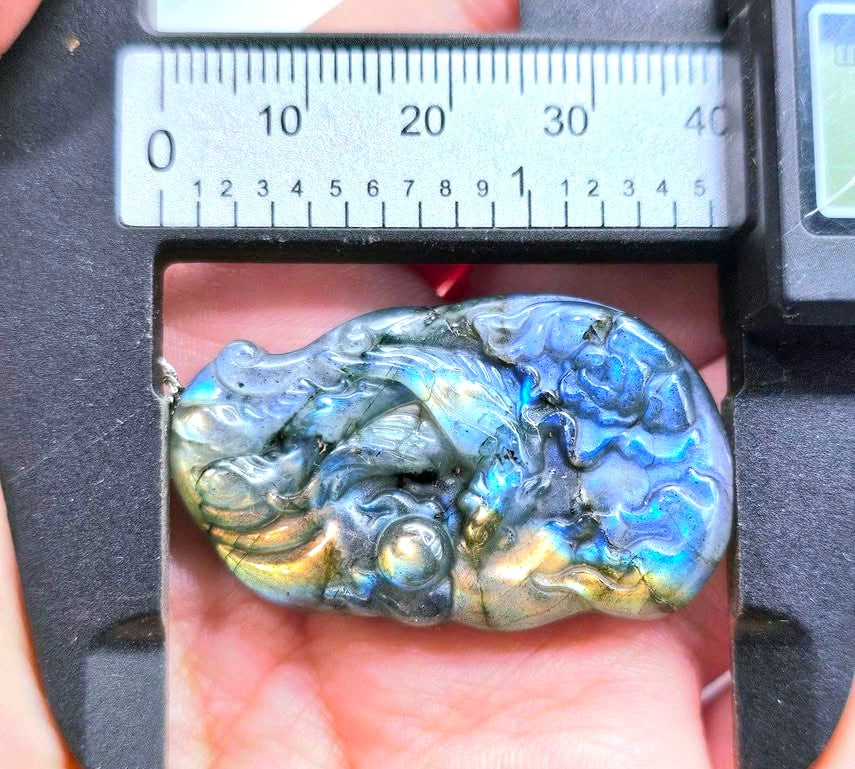 A polished iridescent labradorite stone pendant held in a caliper over a person's hand, showing blue, green, and gold flashes.