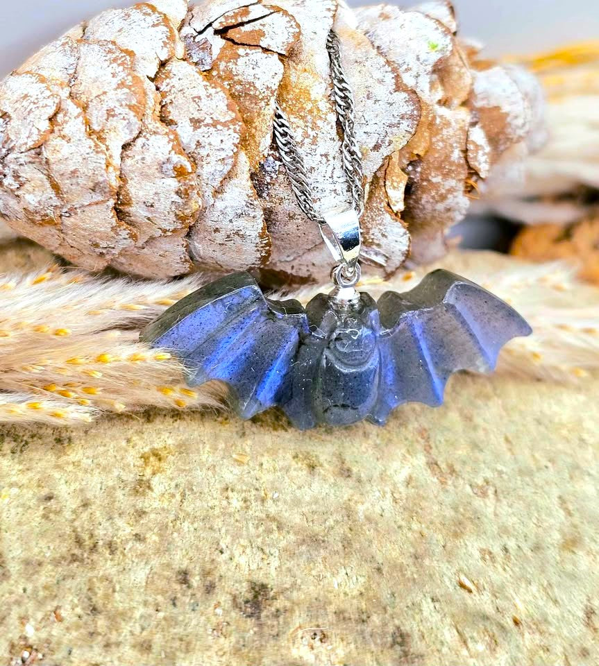 A silver bat pendant necklace with a translucent blue glass or resin wing design, displayed on a textured rock with a pinecone and dried grass.