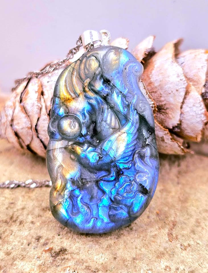 A carved labradorite pendant necklace featuring a detailed phoenix design, resting on a textured stone surface with a pine cone prop. The pendant displays a strong blue, green, and gold iridescent play-of-color.