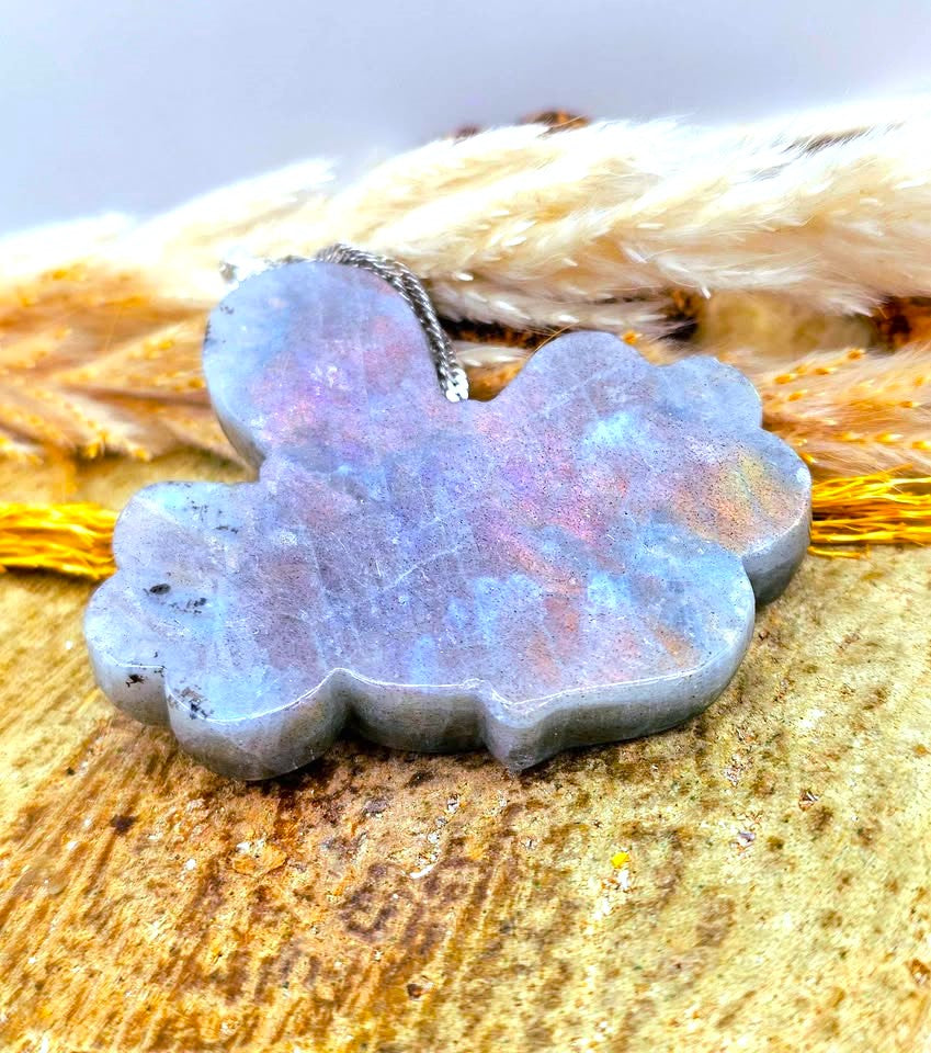 A cloud-shaped labradorite stone pendant with a silver bail, resting on a textured wooden surface with dried grass in the background.