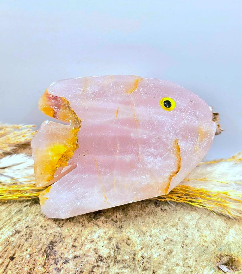 A pink rose quartz crystal fish charm with a yellow and black eye, resting on a textured beige surface with dried yellow grass against a light gray background.