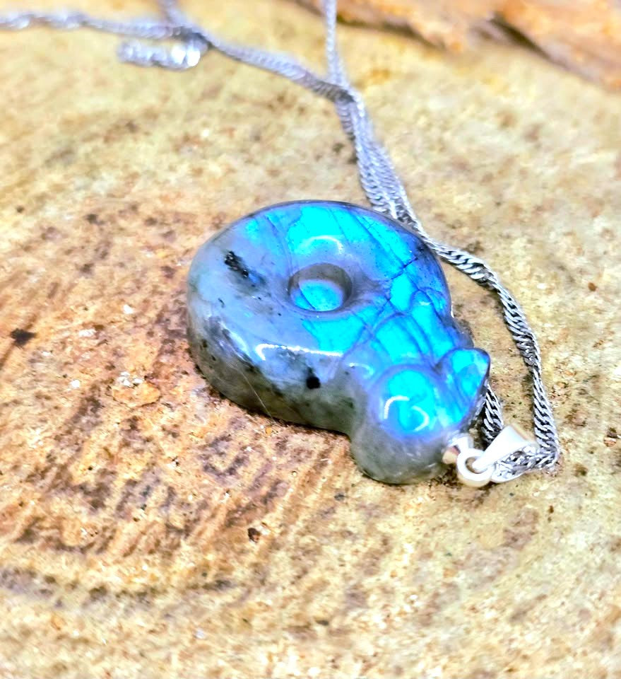 A silver necklace with a polished Labrador cat pendant displaying vibrant blue and green iridescent opalization, resting on a textured stone surface.