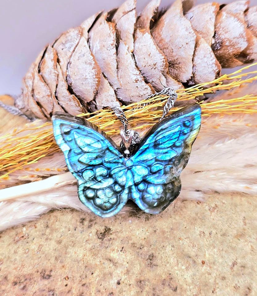 A butterfly-shaped pendant necklace with iridescent blue and green wings, resting on dried grass in front of a pinecone on a sandy surface.