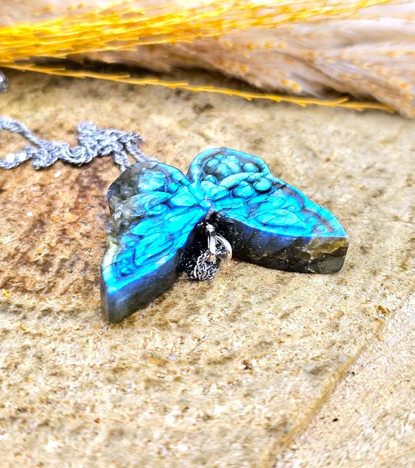A butterfly-shaped pendant necklace made from iridescent labradorite stone with a silver chain, resting on a textured stone surface with dried grass in the background.