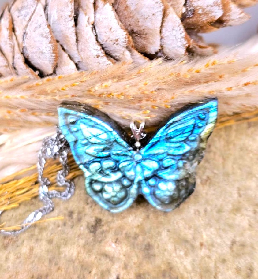 A butterfly-shaped pendant necklace with iridescent blue and green wings, resting on dried grass and a pinecone. The silver-toned chain is visible on the left.