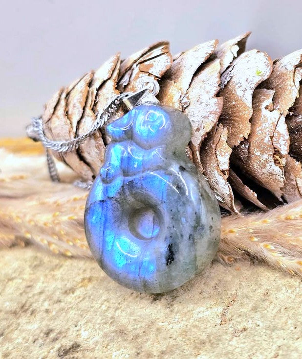 A polished labradorite pendant necklace with a silver-toned chain, resting in front of a pine cone and dried grass on a sandy surface.