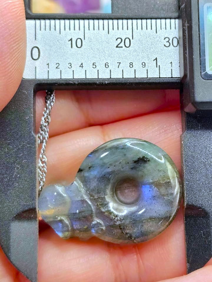 A polished labradorite pendant shaped like a teardrop or hook, resting on a person's fingers and measured under a digital caliper with a ruler scale visible above it. The pendant has a silver chain and displays natural labradorite coloration with gray, blue, and purple iridescence.