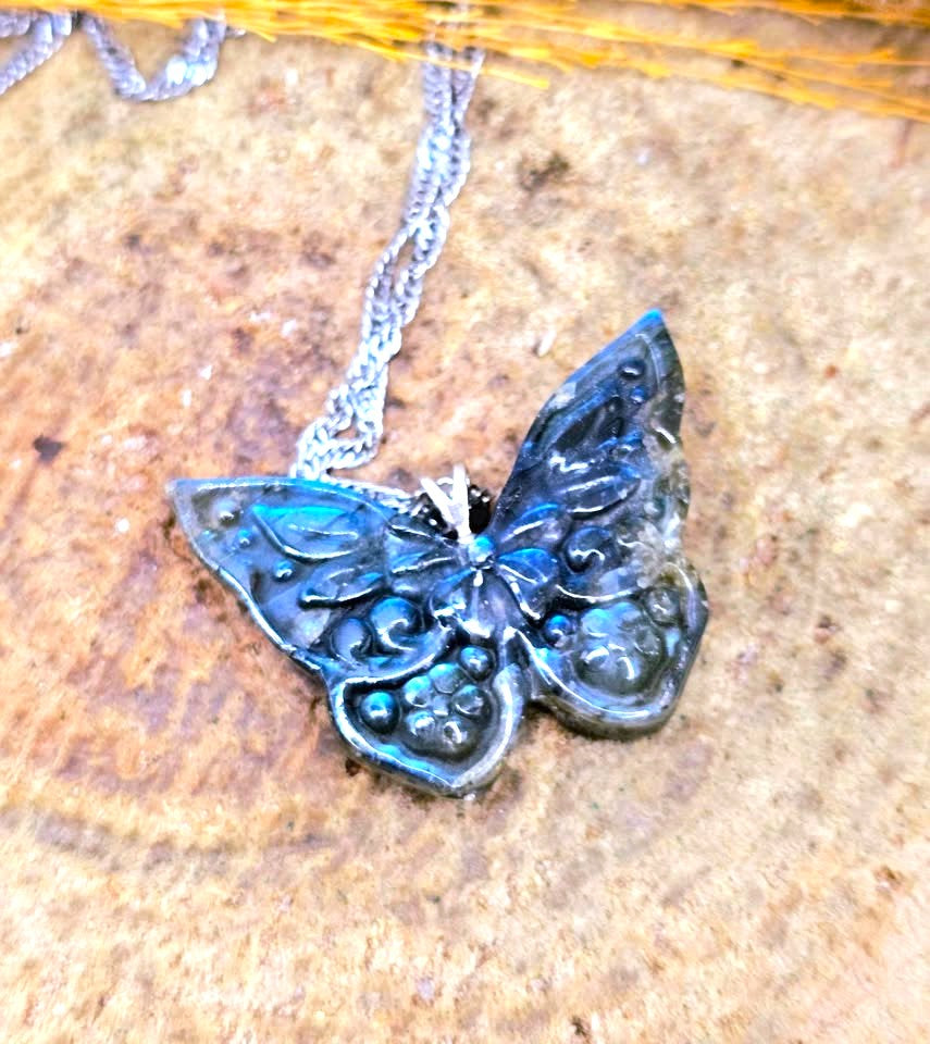 A butterfly-shaped pendant necklace with a silver-toned chain, featuring dark blue and black enamel detailing with small blue stones, resting on a textured brown surface.