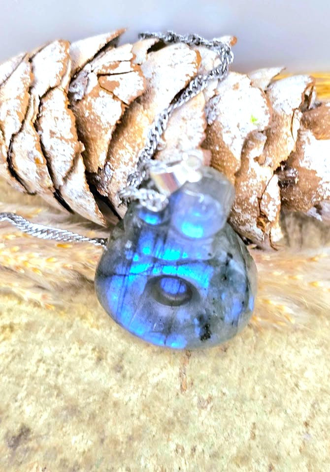 A labradorite pendant necklace with a silver chain, resting on a pinecone and dried grass. The pendant displays a strong blue iridescent labradorescent sheen.