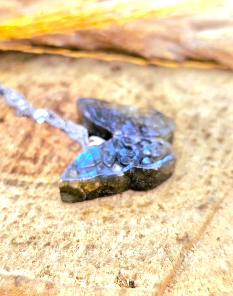 A close-up of a carved labradorite pendant necklace resting on a textured stone surface. The pendant features a butterfly design with a dark gray and black base that glows with blue and green iridescent colors, suspended from a silver-toned chain.