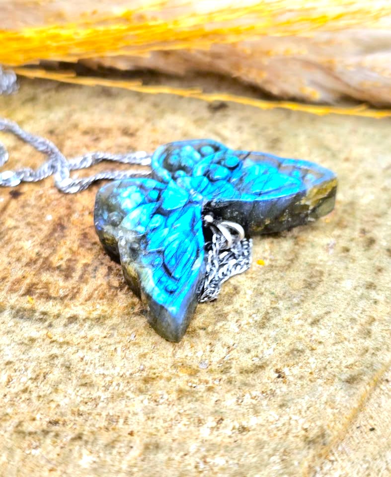 A close-up of a butterfly-shaped pendant made from a polished labradorite stone showing vibrant blue iridescence, hanging from a silver chain on a textured stone surface.