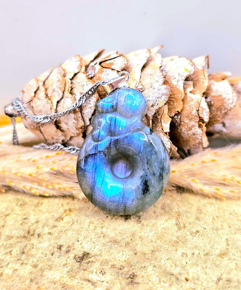A carved labradorite pendant necklace with a silver-toned chain, displayed on a sandy surface with a pine cone and dried grass in the background.