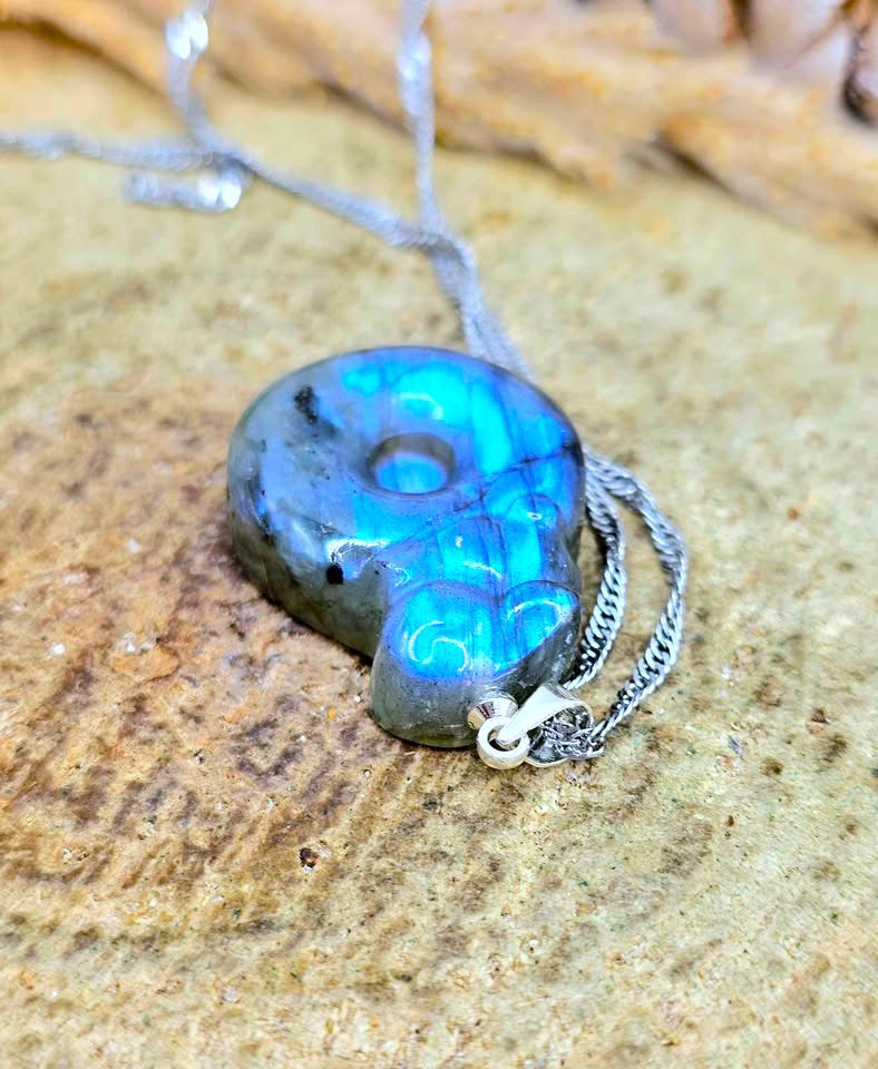 A polished labradorite gemstone pendant necklace with a silver chain, resting on a textured sandy surface. The pendant displays a vibrant blue and green iridescent play-of-color with dark gray matrix lines.