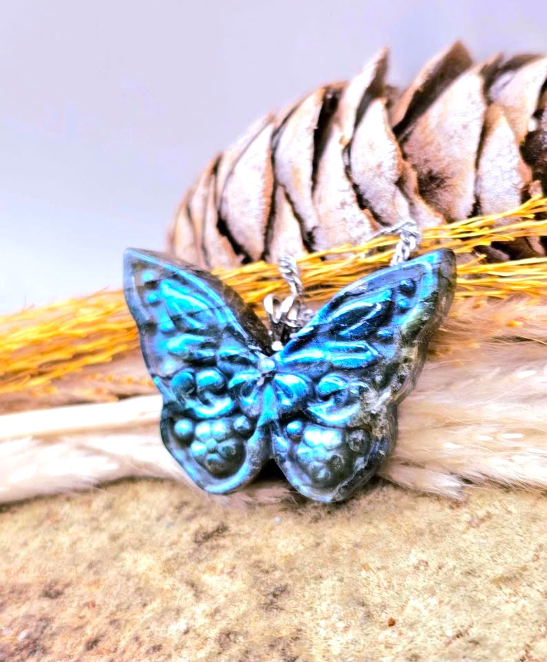 A butterfly-shaped pendant with iridescent blue and teal enamel, featuring raised scrollwork details and a silver-tone bail, displayed on natural brown and beige textured props.